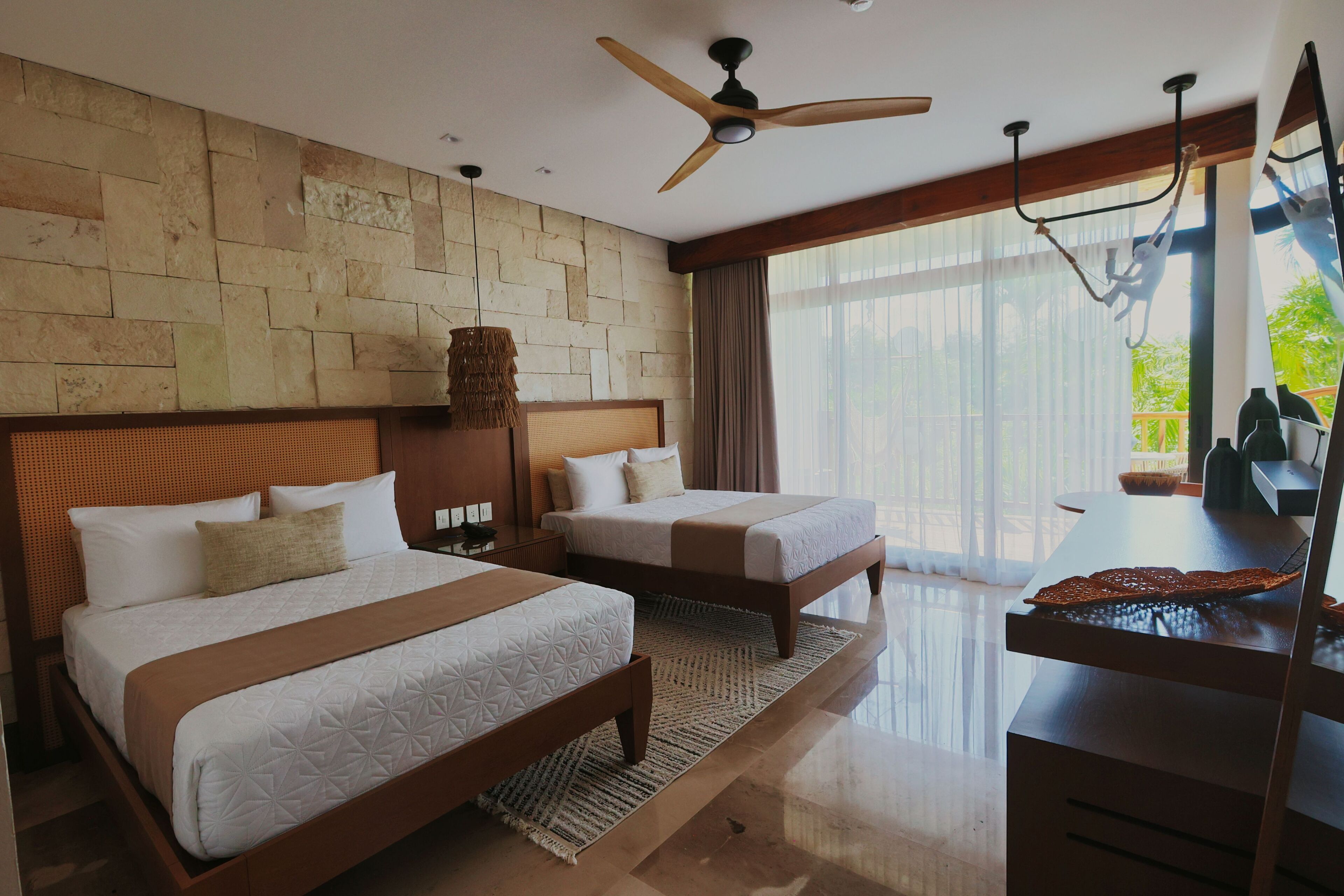 A hotel room with two beds, a desk, a ceiling fan, and a large window with a view of greenery.