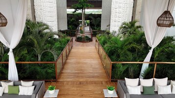 Un sendero de madera que conduce a un área central con una estructura circular, rodeada de exuberante follaje y cortinas blancas.
