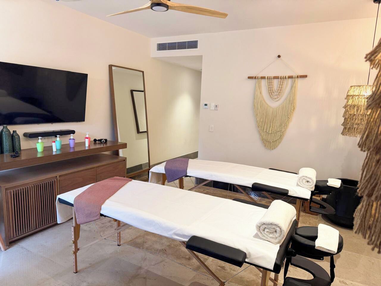 A room with two massage tables, a television, a wooden cabinet with toiletries, and a wall-mounted macrame piece.