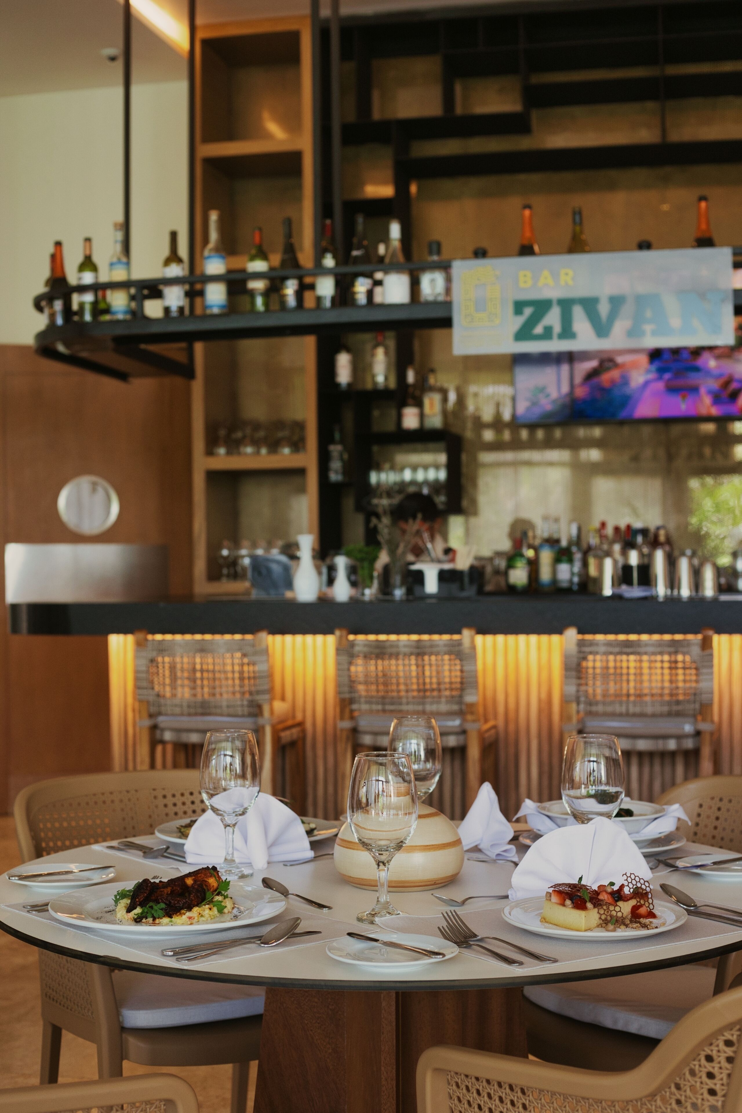 Un ambiente de restaurante con mesas preparadas para comer, una zona de bar con botellas, y un letrero que dice "BAR ZIVAN".