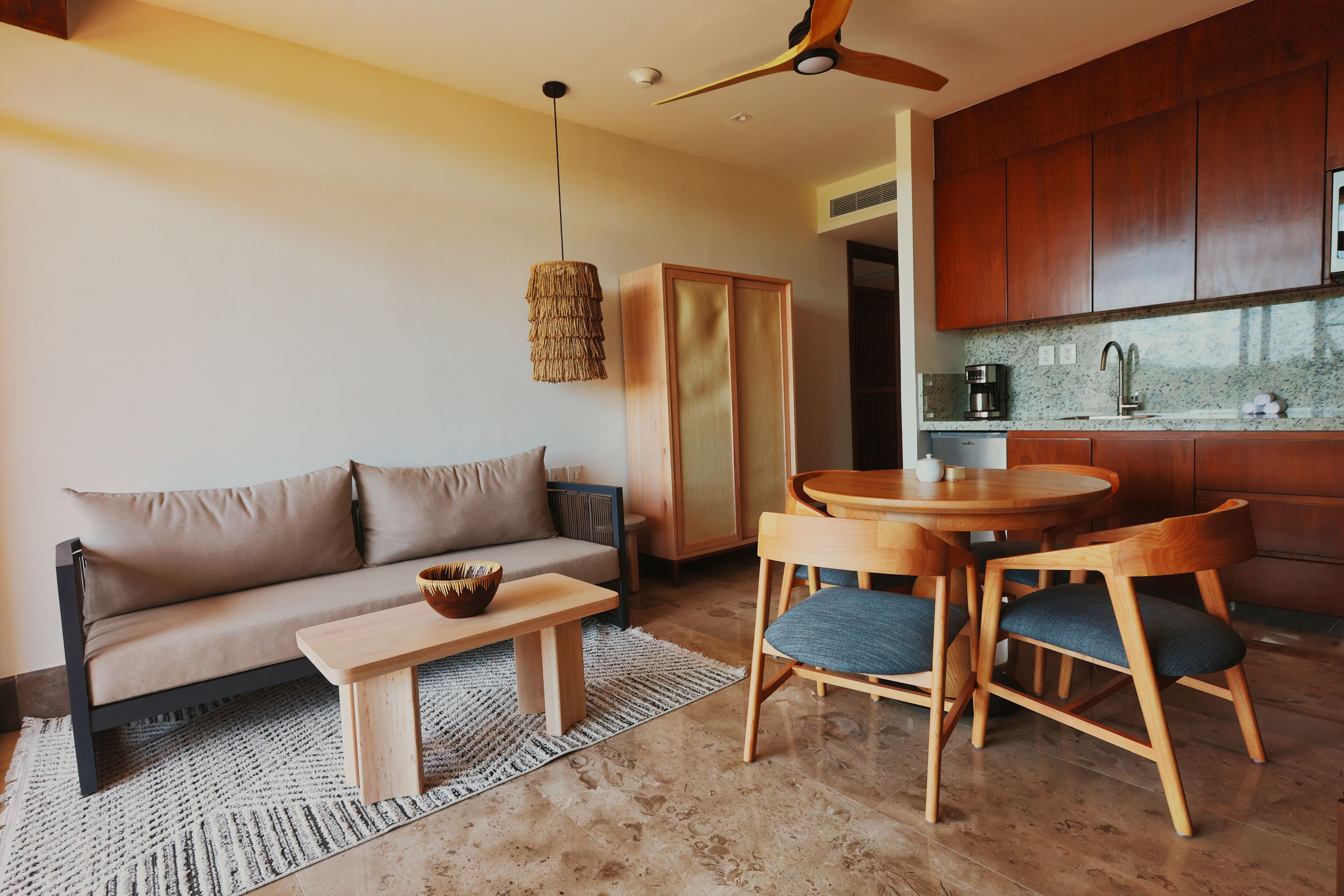 Una sala moderna con sofá, mesa de centro y sillas, una zona de cocina con armarios de madera y ventilador de techo, además de una alfombra con textura.