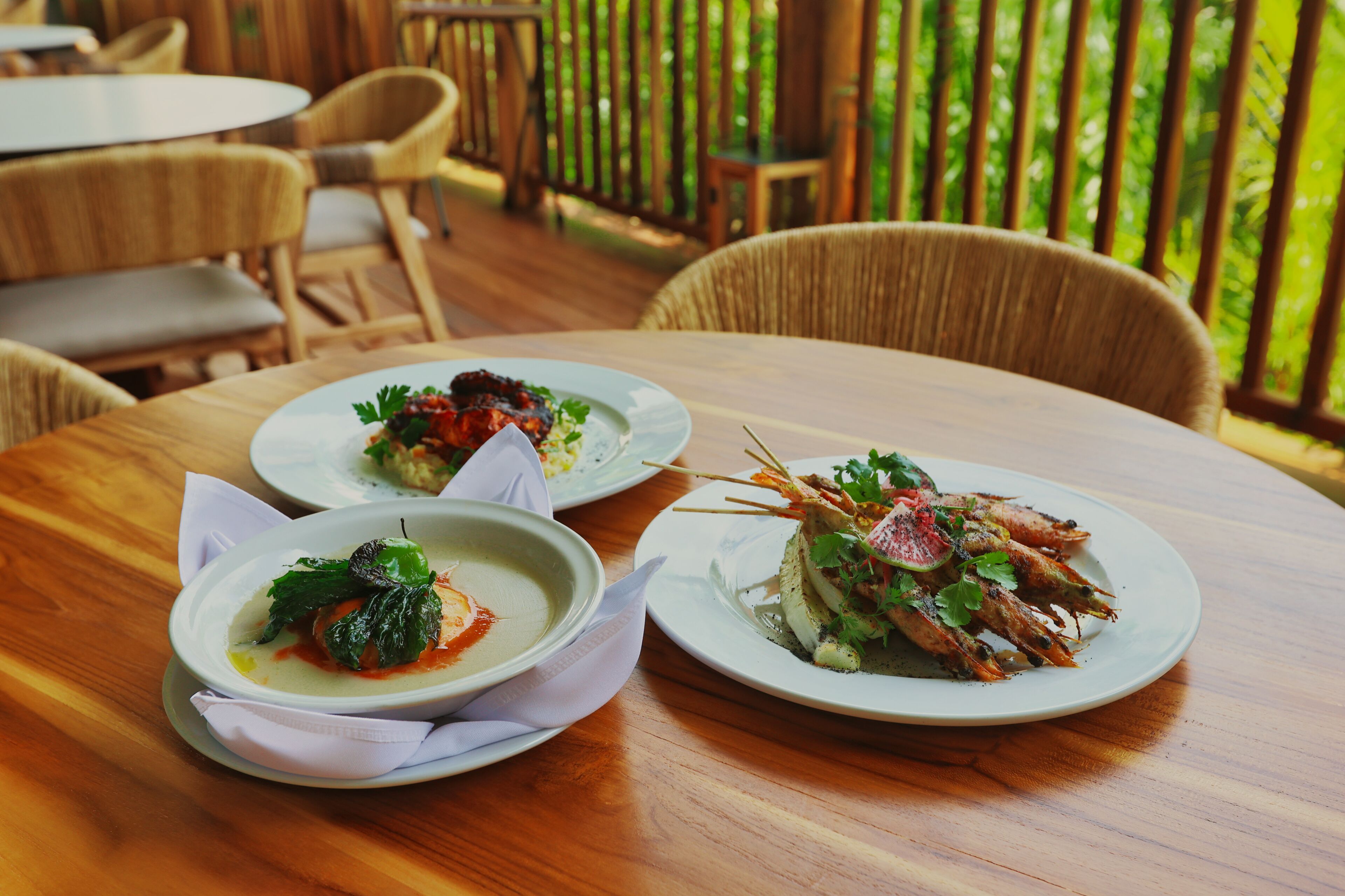 Two plates of food on a wooden table in an outdoor dining area with wicker chairs and a wooden deck.