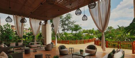 A covered outdoor seating area with sofas, chairs, and tables, surrounded by greenery and a wooden deck.