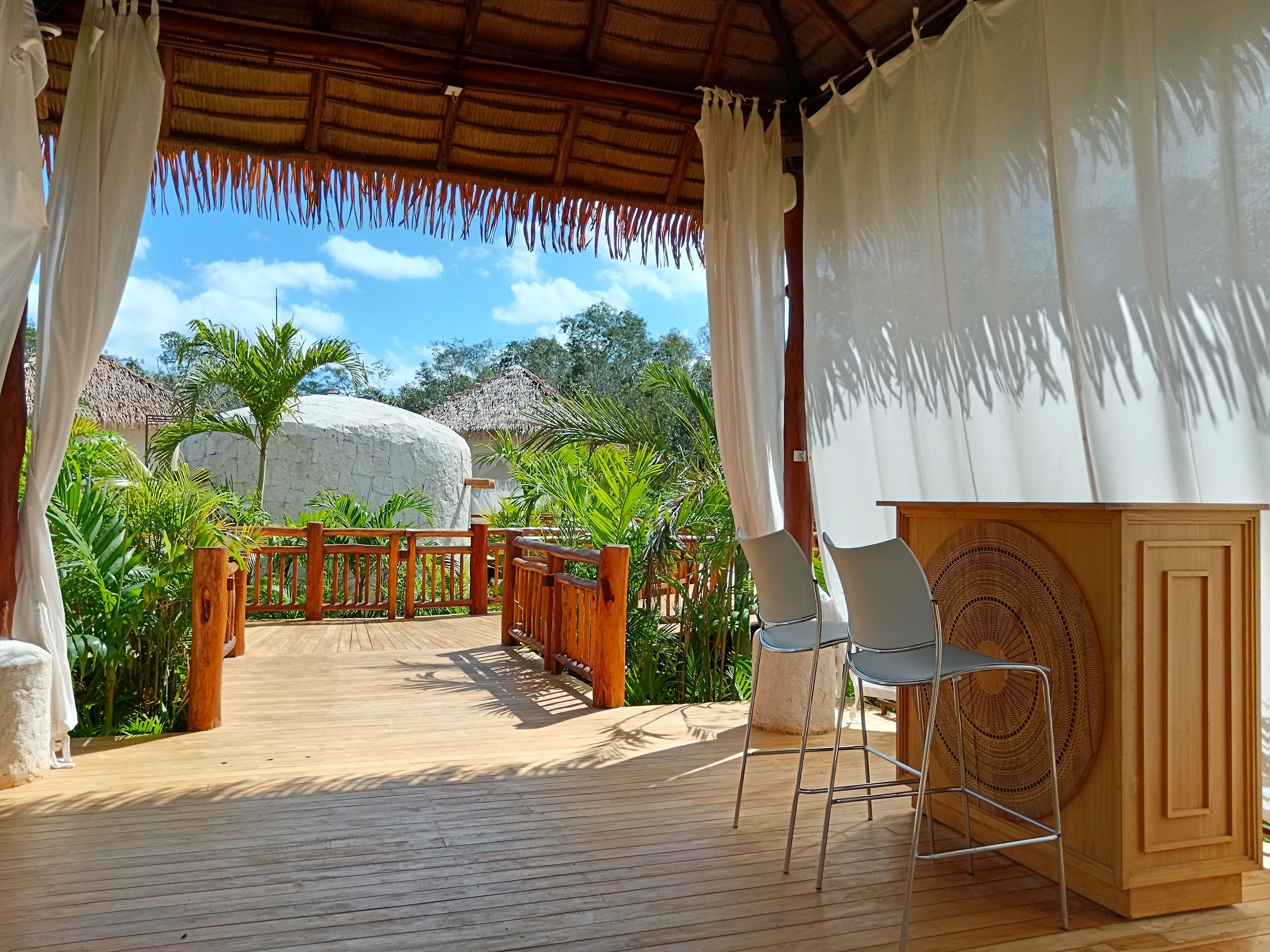 A wooden deck with a thatched roof, white curtains, and a view of a large rock and greenery.