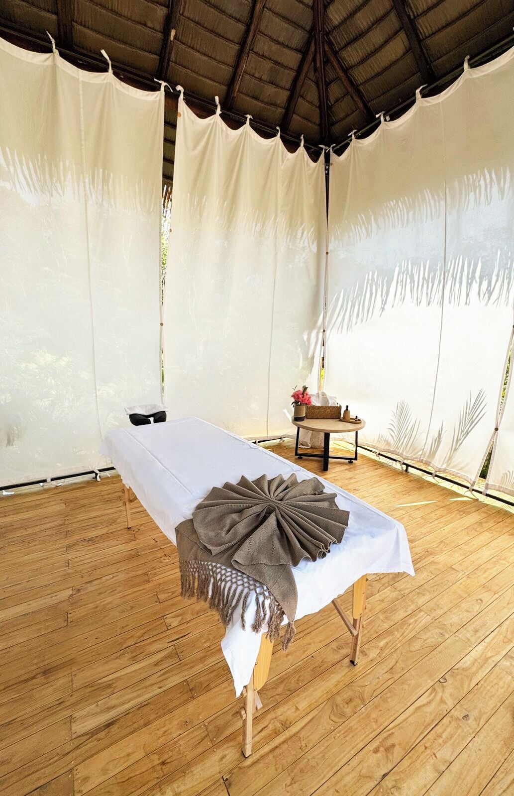 Un área con carpas, un suelo de madera, una mesa con mantel blanco y un paño marrón decorativo, y una pequeña mesa redonda con un florero y una cesta.