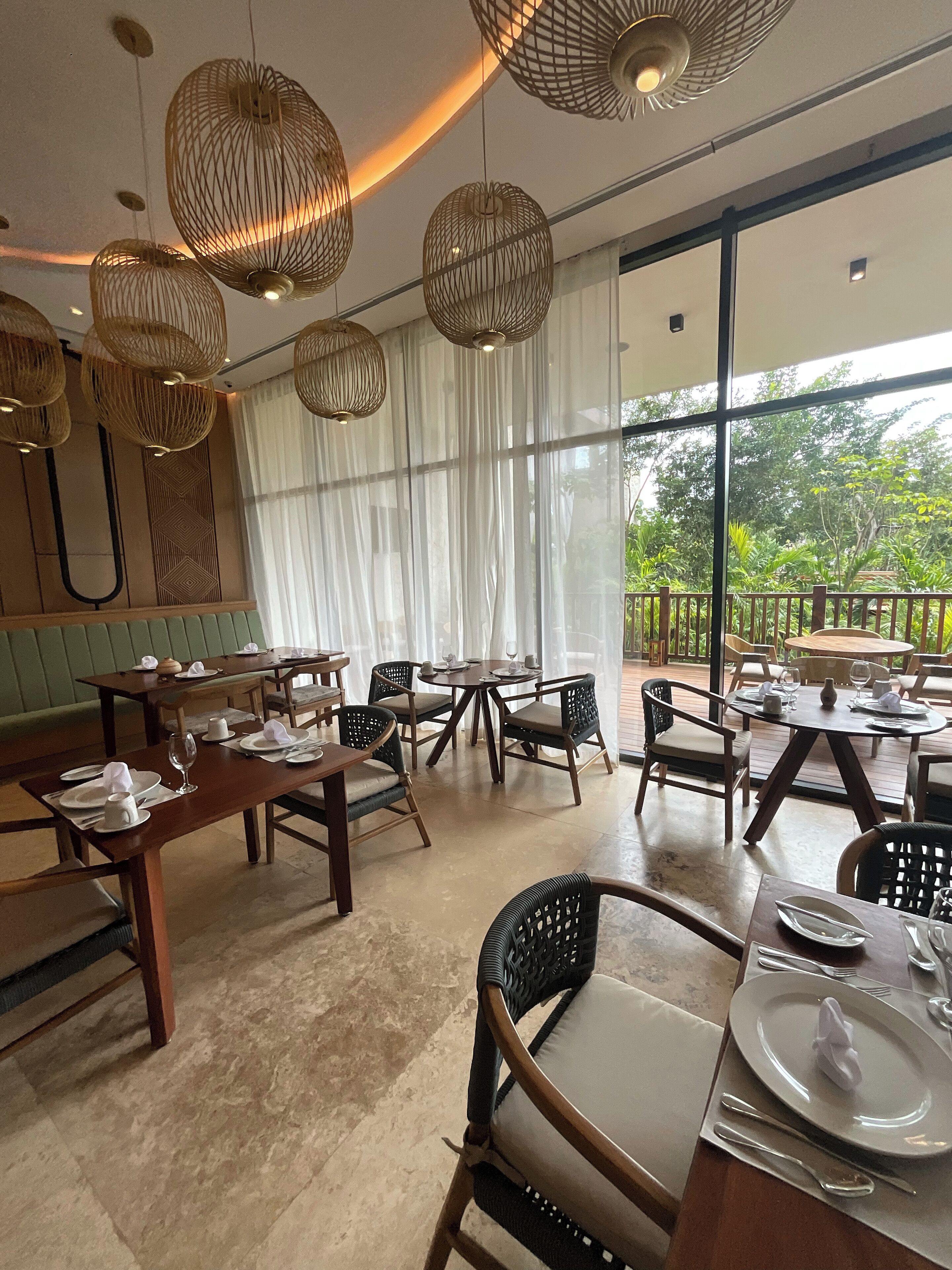 A restaurant with wooden tables and chairs, large windows with a view of greenery, and hanging wicker light fixtures.