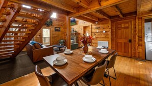 A wooden cabin interior with a dining table set for four, a sofa, a staircase, and a kitchen area. - Cozy Cabin Retreat: Hot Tub, Fire Pit & Game Room – Perfect Getaway! (Sevierville)