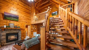 A log cabin interior with a fireplace, wooden staircase, and a cozy seating area. - Cozy Cabin Retreat: Hot Tub, Fire Pit & Game Room – Perfect Getaway! (Sevierville)