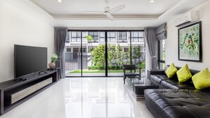 Een moderne woonkamer met een flatscreen televisie, een zwarte leren bank met groene kussens, een salontafel en een grote schuifpui die uitkomt op een balkon met beplanting.