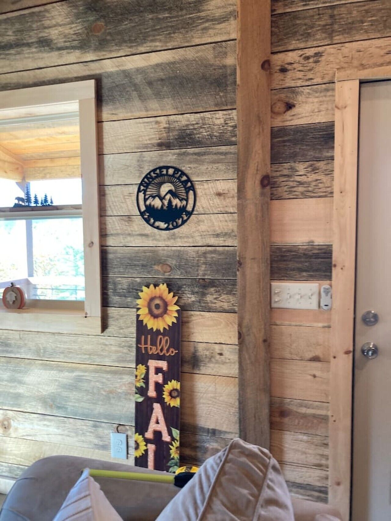 A rustic cabin interior with a wooden wall, a circular emblem, and a vertical sign with the word 'Hello' and sunflowers.