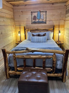Ein Schlafzimmer einer Holzhütte mit einem Bett, zwei Nachttischlampen, einer Bank und einem Baumgemälde an der Wand.