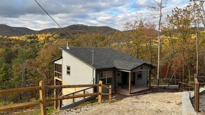 Une cabane à deux étages dotée d’une terrasse en bois, entourée d’une clôture en bois, et se dressant sur un fond de feuillage automnal et de collines.