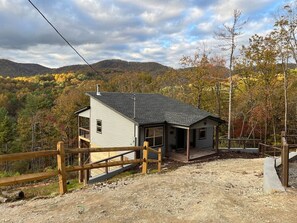 Eine zweistöckige Holzhütte mit einer Holzterrasse, umgeben von einem Holzzaun, vor dem Hintergrund von herbstlich gefärbten Bäumen und Hügeln.