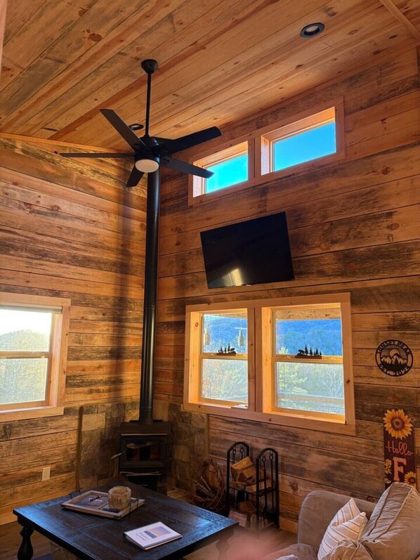 Ein gemütliches Blockhaus-Interieur mit Holzvertäfelung, Deckenventilator, Kamin, Wand-Fernseher und Fenstern mit Blick ins Freie.