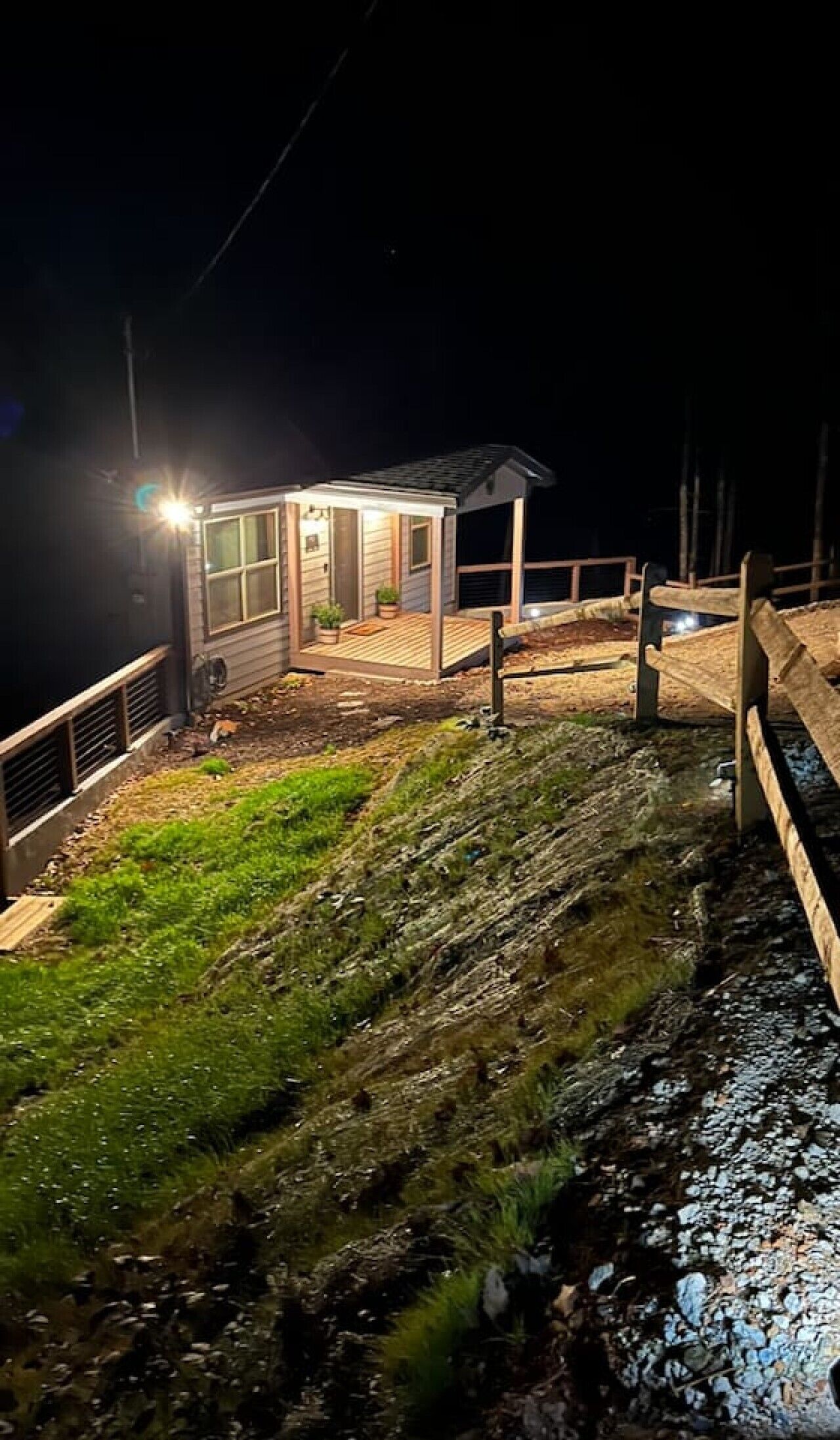 A small, illuminated structure with a wooden deck, situated on a sloped terrain with a metal railing.