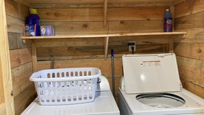 Un espace buanderie comprenant un lave-linge et un sèche-linge, un panier à linge blanc et des étagères contenant des produits de nettoyage.