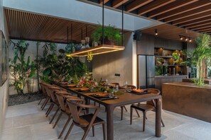 A modern dining area with a long wooden table, multiple chairs, and a variety of plants. - Villa Baratega (Kerobokan)
