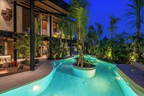 A swimming pool with a curved design, surrounded by tropical plants and a modern building with a wooden exterior. - Villa Baratega (Kerobokan)