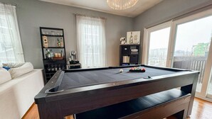 A pool table in a modern living room with a sofa, a bookshelf, and a view of the city through large windows. - Southie Condo - Near Subway - Bring Pets - Pool Table (Boston)