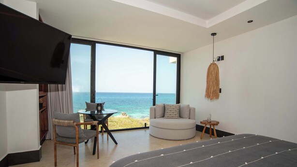 A bedroom with a large bed, a TV, a chair, a small table, and a view of the ocean.