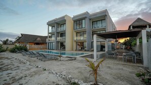 Un hôtel moderne à plusieurs étages, doté d’une piscine, d’un espace extérieur aménagé et d’une structure surmontée d’un toit de chaume.