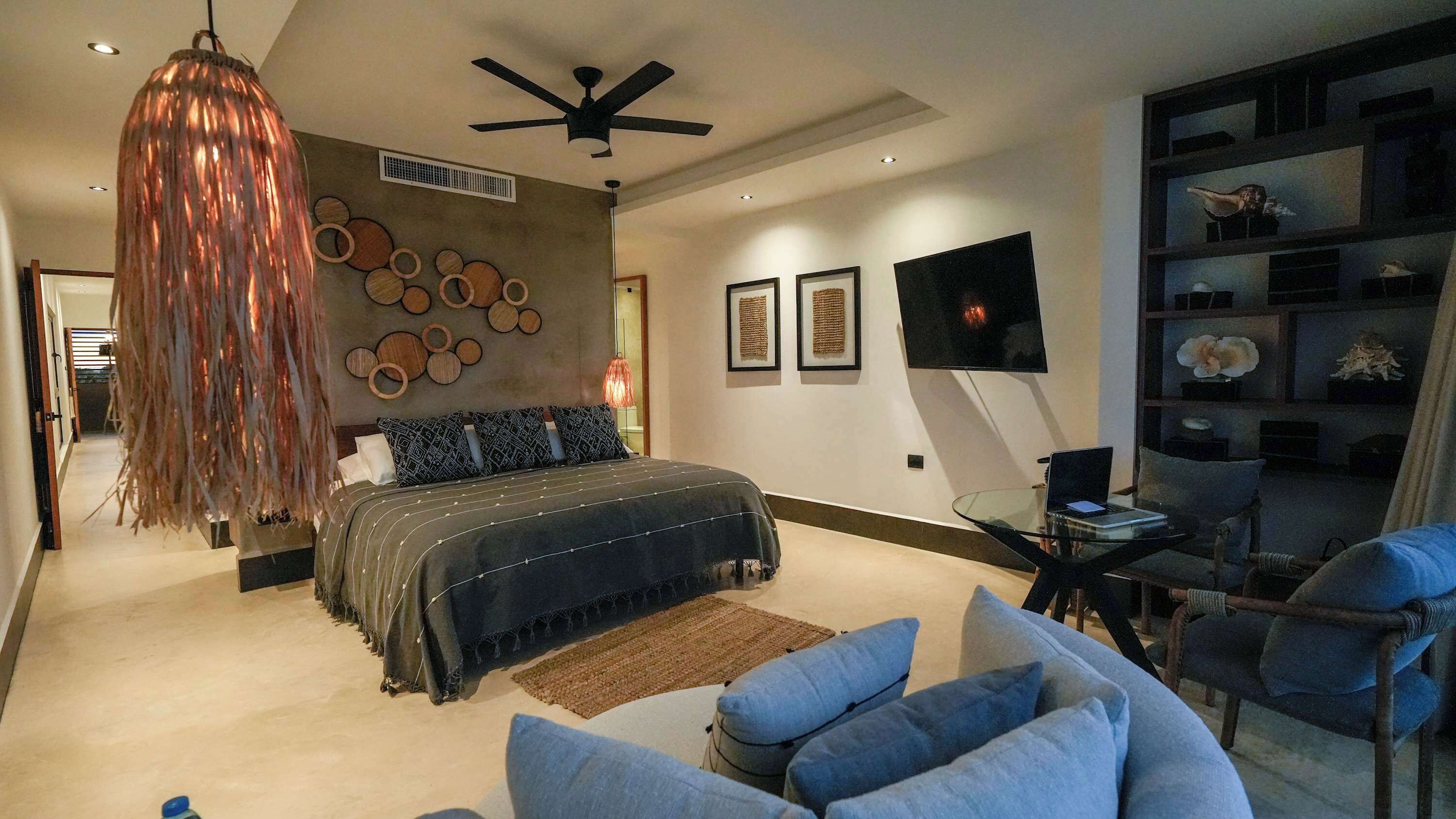 A modern bedroom with a large bed, a ceiling fan, a glass desk, and a wall-mounted TV.