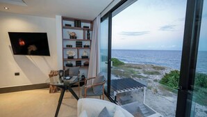 Ett modernt vardagsrum med ett stort fönster med utsikt över en strand, en platt-TV som är monterad på väggen och ett glasbord med en bärbar dator och en stol.