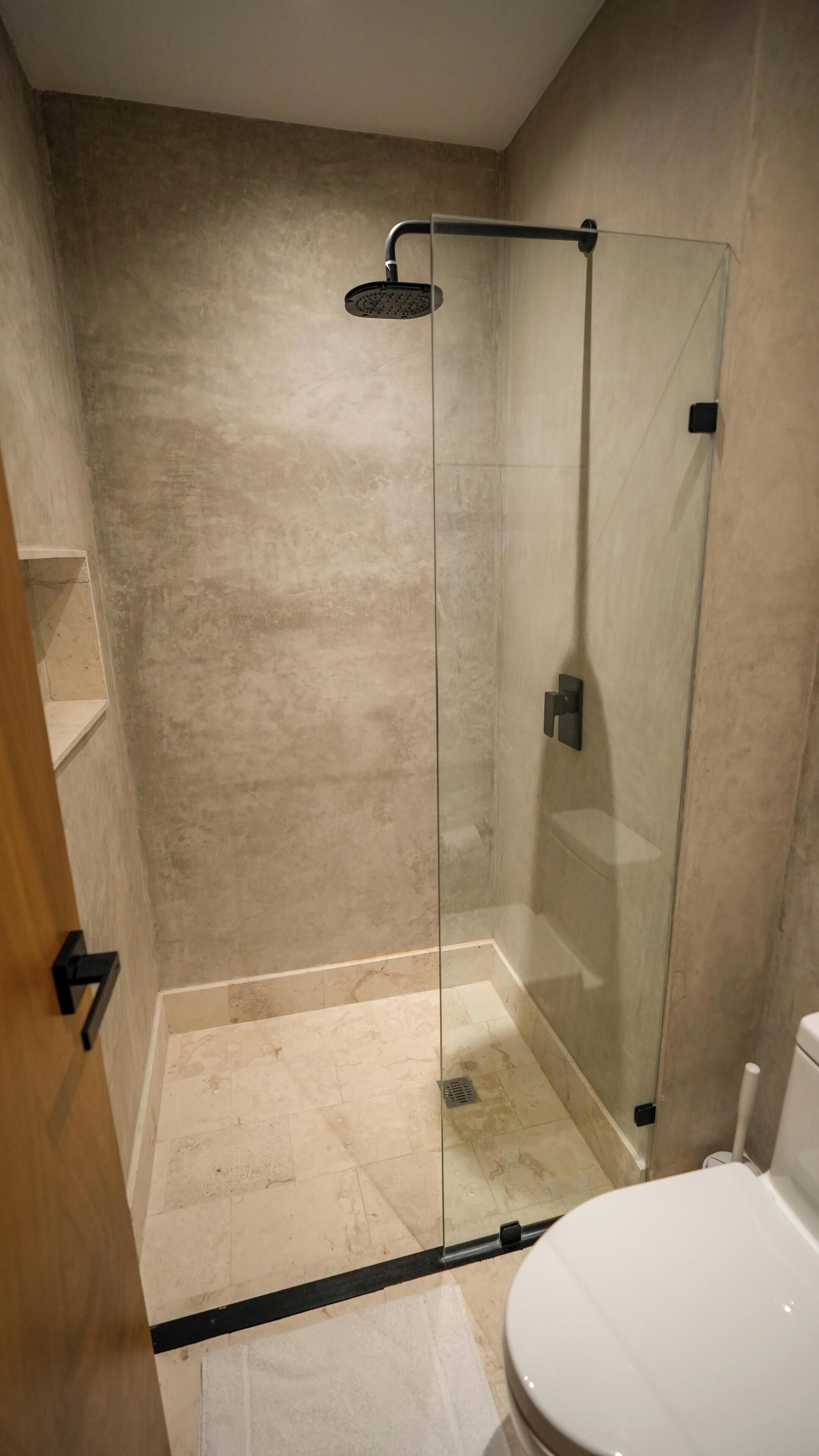 Et glasvægshavn med brusehoved i hånden, et hvidt toilet og et beige klinkegulv.