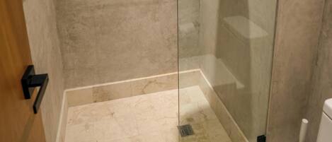 Et glasvægshavn med brusehoved i hånden, et hvidt toilet og et beige klinkegulv.