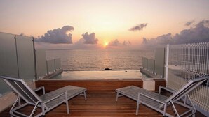 En terrass med två solstolar som vetter mot en pool och havet, med utsikt över solnedgången.