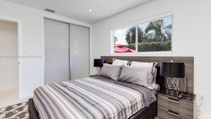 Une chambre à coucher avec un lit, une table de chevet, une lampe et une fenêtre donnant sur des palmiers.