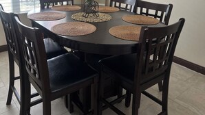 Un coin repas avec une table ronde, des chaises en bois foncé, des sets de table en fibres tressées, un lustre, un tableau encadré au mur et une fenêtre avec des stores.