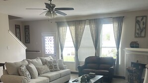 Un salon comprenant un canapé, deux fauteuils, une table basse, un pouf noir, un ventilateur de plafond et une cheminée.