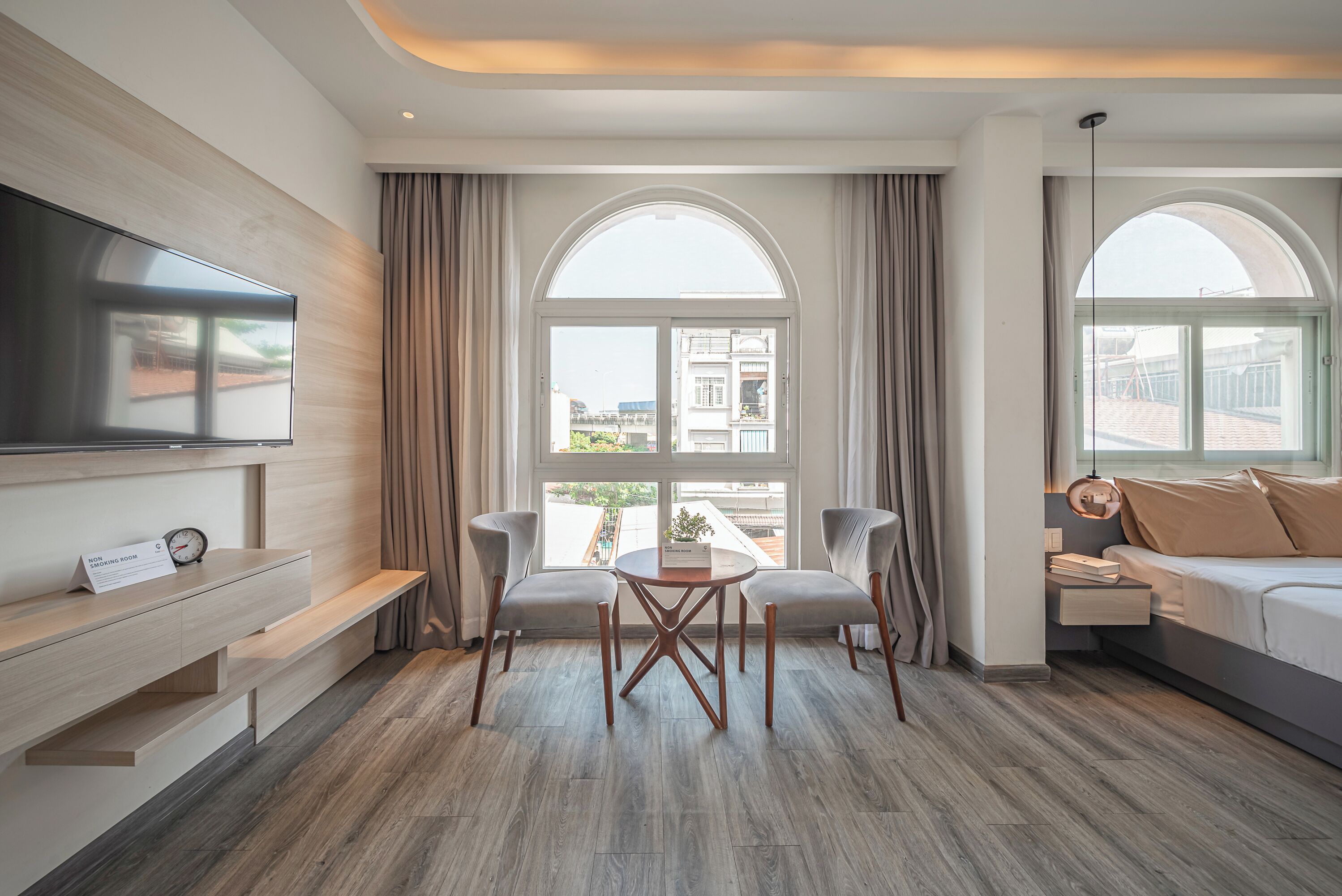 A modern hotel room with a large TV, a dining table with chairs, a sofa, and a large arched window offering a city view.