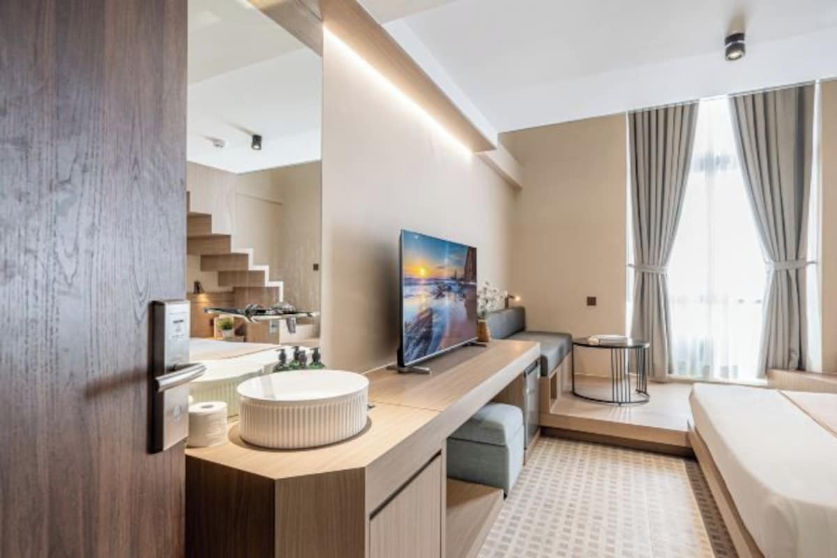 Habitación de hotel moderna con tocador de madera, un espejo grande, un televisor de pantalla plana, cama y ventana con cortinas.