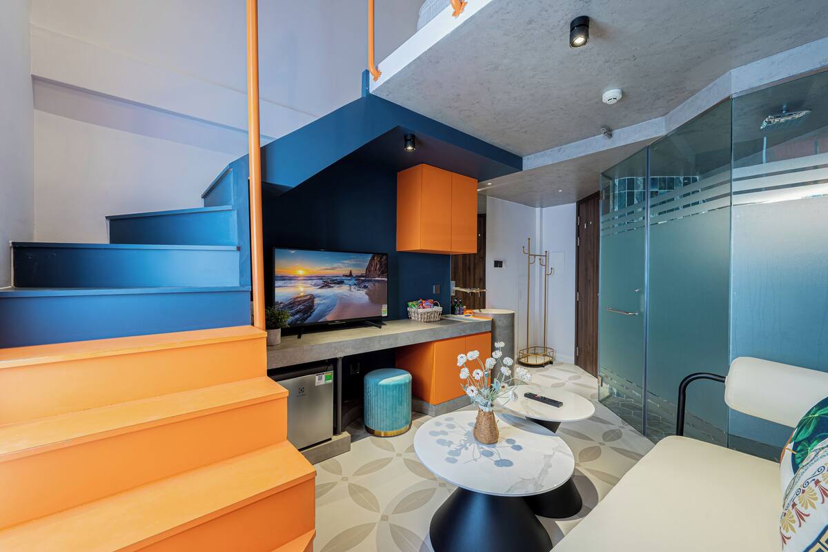 A modern living room with a large flat-screen TV, orange and blue furniture, a glass partition, and a staircase.
