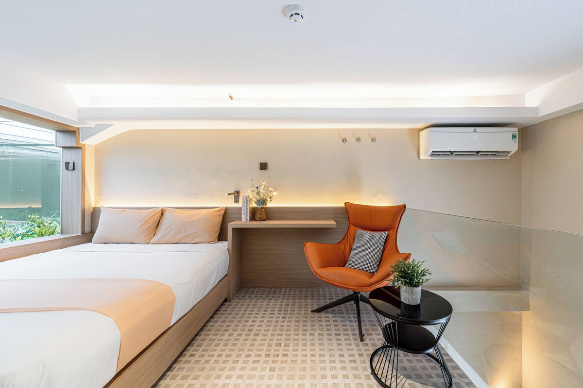 A modern hotel room with a large bed, a small table with a chair, a potted plant, and a built-in air conditioning unit.
