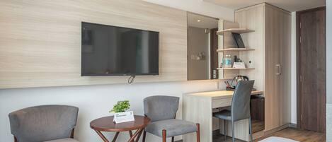 A modern hotel room with a flat-screen TV mounted on the wall, two grey armchairs, a small round table with a plant, a desk with a chair, and built-in shelving with toiletries.