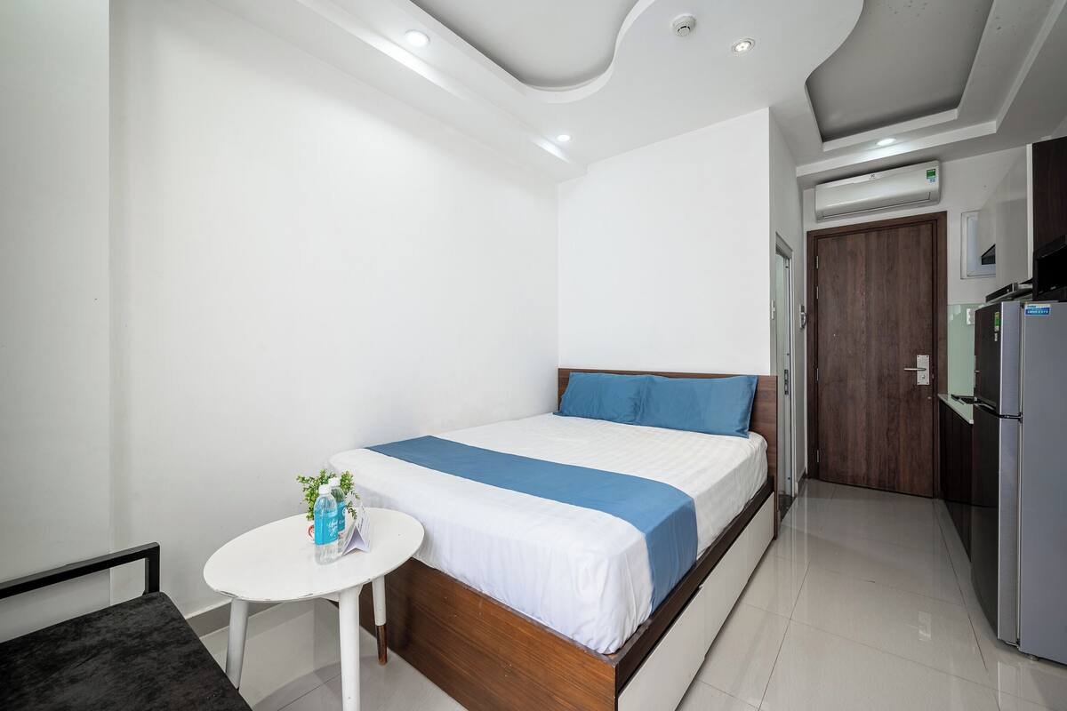 A modern hotel room with a neatly made bed, a small round table with a plant, and a kitchenette area.