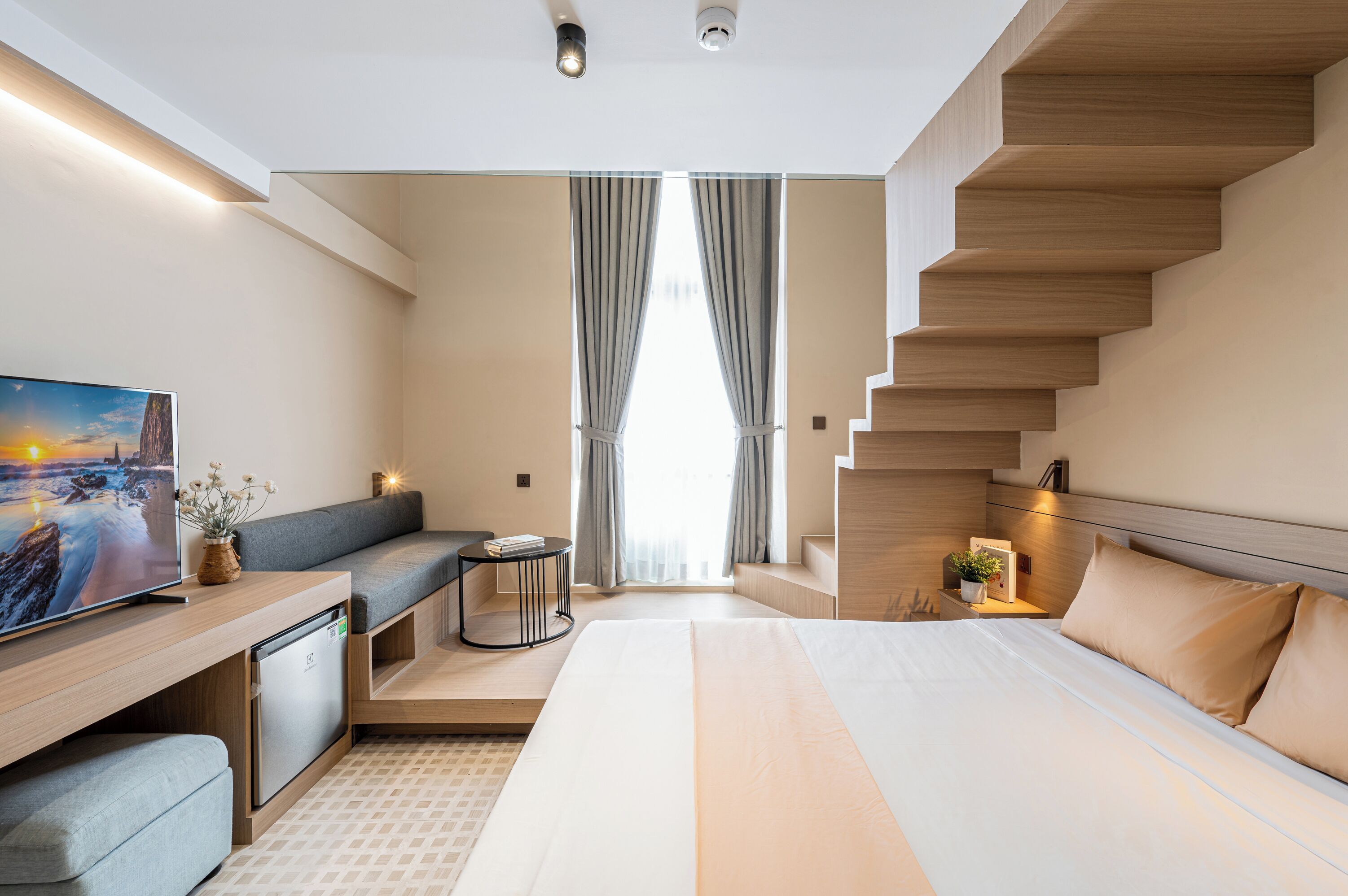 A modern hotel room with a bed, a seating area, a TV, and a staircase.