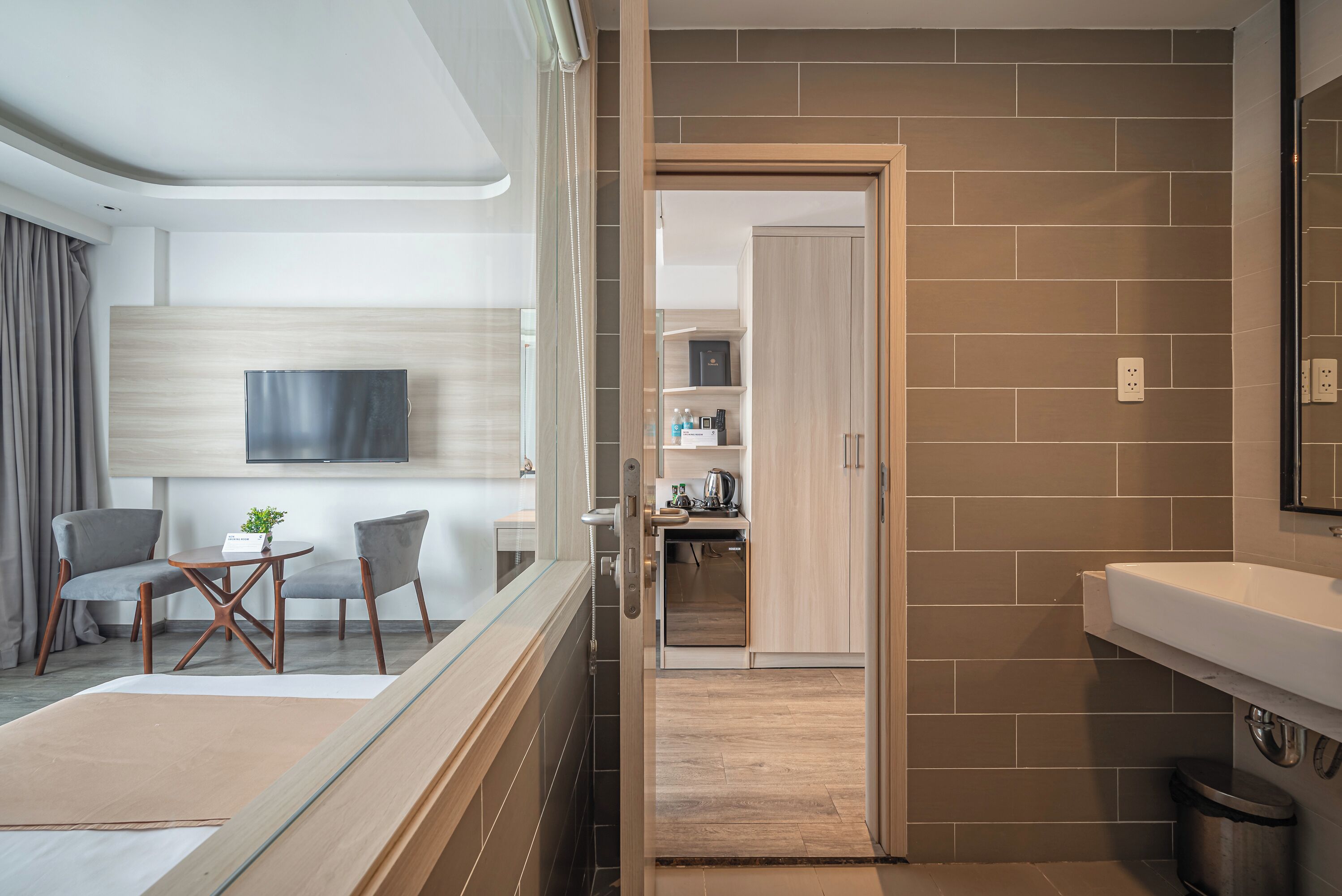 A modern hotel room with a seating area, a TV mounted on the wall, a kitchen area visible through an open door, and a bathroom with a sink and mirror.