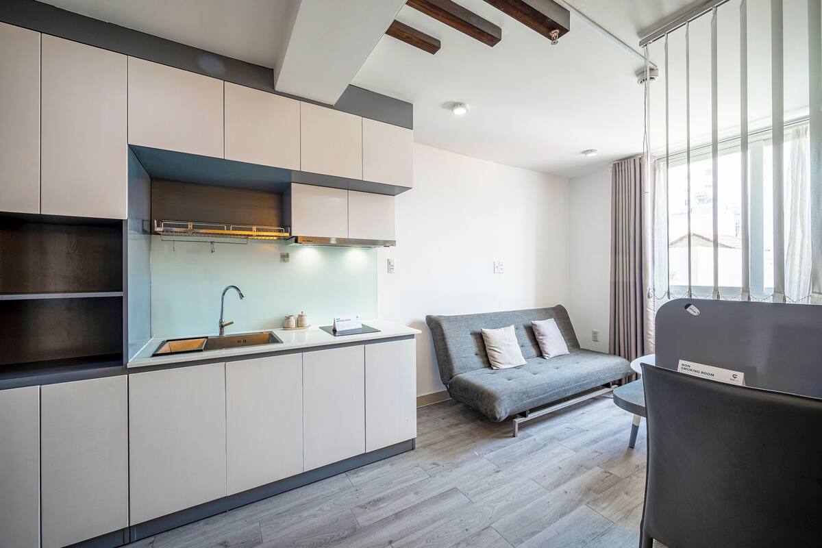 A modern kitchen with built-in cabinets, a sink, and a stove. There is a grey sofa with cushions and a small table with a chair in the living area. A sign on the sofa indicates it is a non-booking room.
