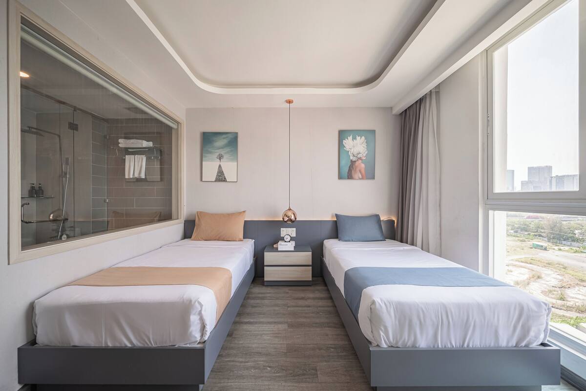 A modern hotel room with two beds, a shower area, and large windows offering a city view.