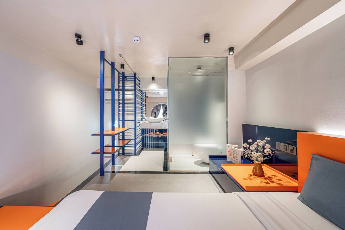 A modern hotel room with a bed, a small table with a vase, a blue and orange wall, and a staircase leading to a bathroom.