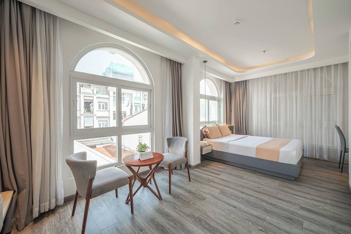 A modern hotel room with a large bed, a seating area with a table, and a view of the city through large windows.