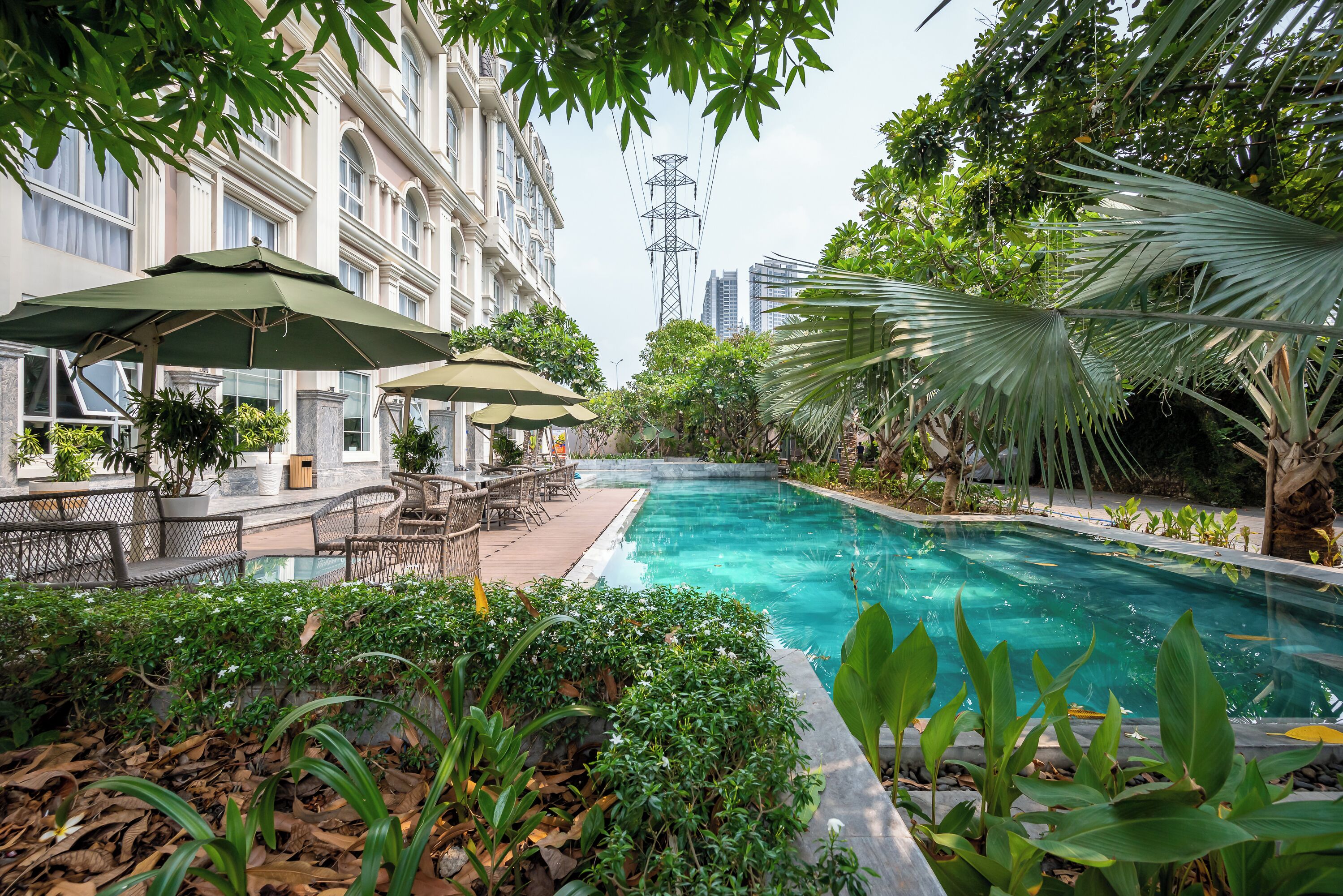 A swimming pool surrounded by lush greenery, outdoor seating with umbrellas, and a view of a tall building in the background.