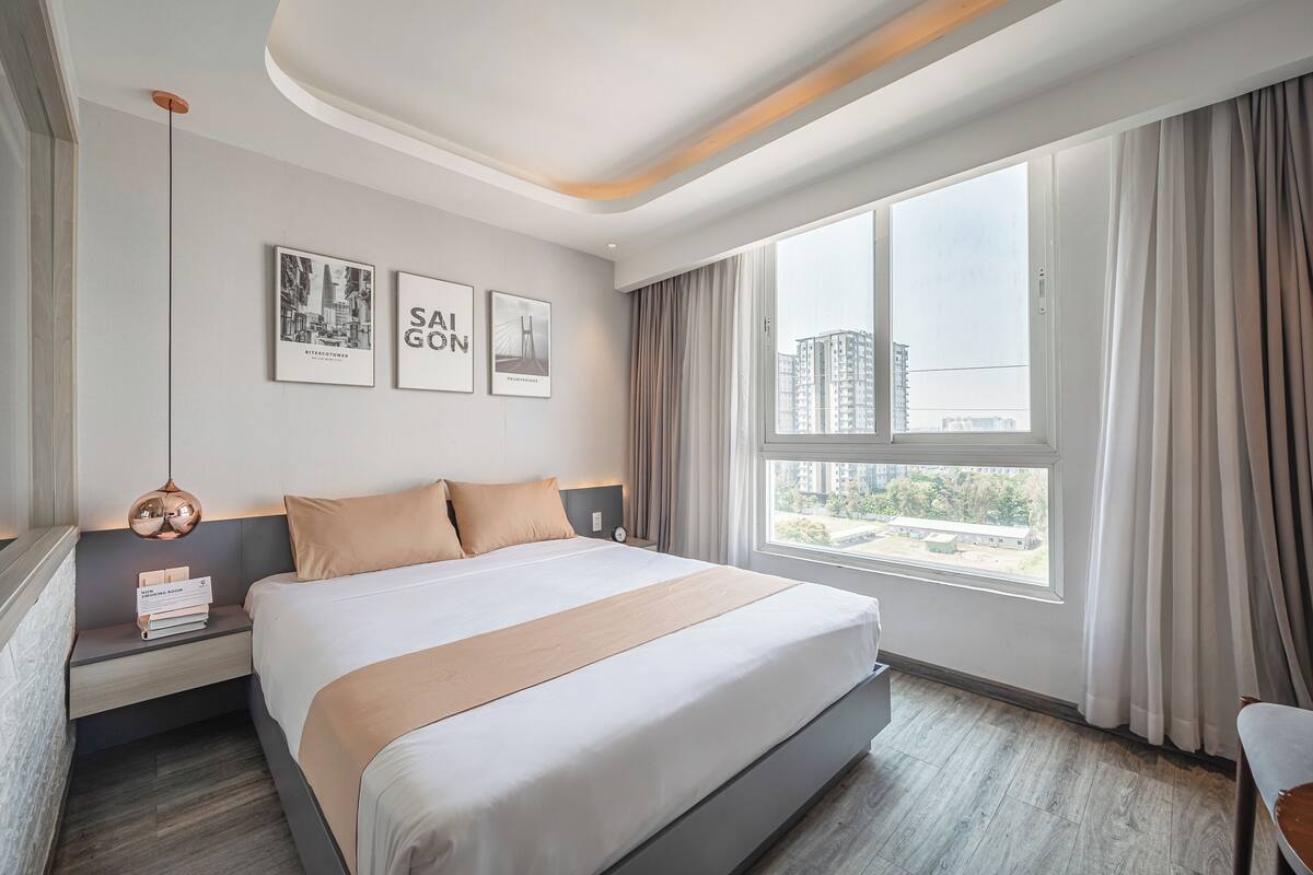 A modern hotel room with a large bed, a nightstand with books, a wall-mounted lamp, and a window with curtains.