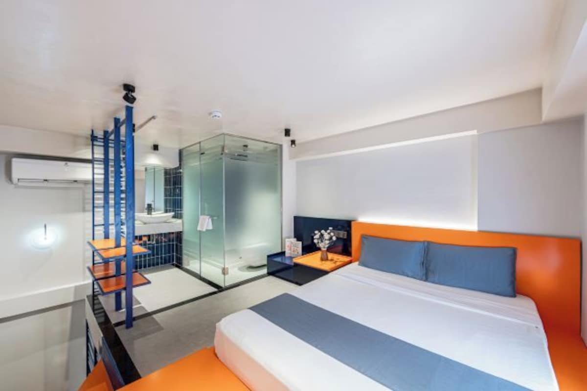Habitación de hotel moderna con una cama grande, cabina de ducha de vidrio, un escritorio pequeño con una silla y un baño con mampara de vidrio.