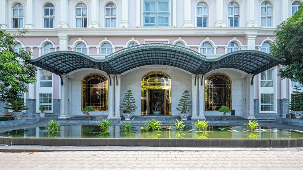 A grand building with a symmetrical facade, a central entrance with a decorative arch, and a surrounding water feature with plants. - Cozrrum Lux - Sonata Imperial (Ho Chi Minh City)
