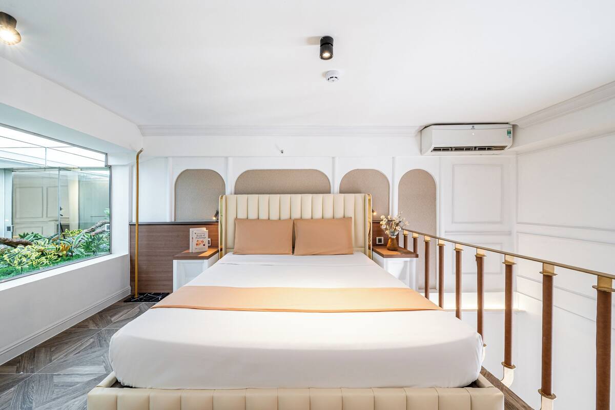 A modern hotel room with a large bed, a view of greenery through the window, and a built-in air conditioning unit.